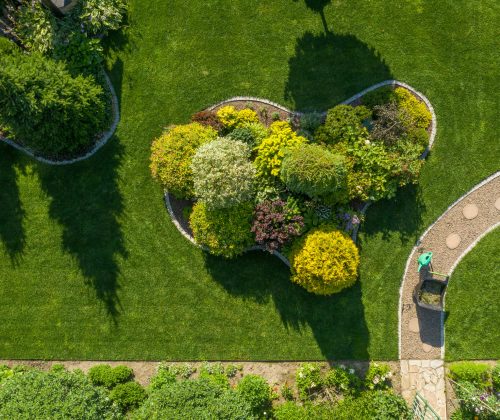 Garden Maintenance and Care Work in Process. Wheelbarrow with Grass Cuttings and a Plants Watering Can Stand on a Twisting Garden Path Next to a Large Landscape Island. Aerial View.