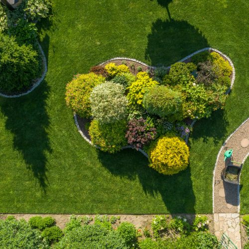 Garden Maintenance and Care Work in Process. Wheelbarrow with Grass Cuttings and a Plants Watering Can Stand on a Twisting Garden Path Next to a Large Landscape Island. Aerial View.